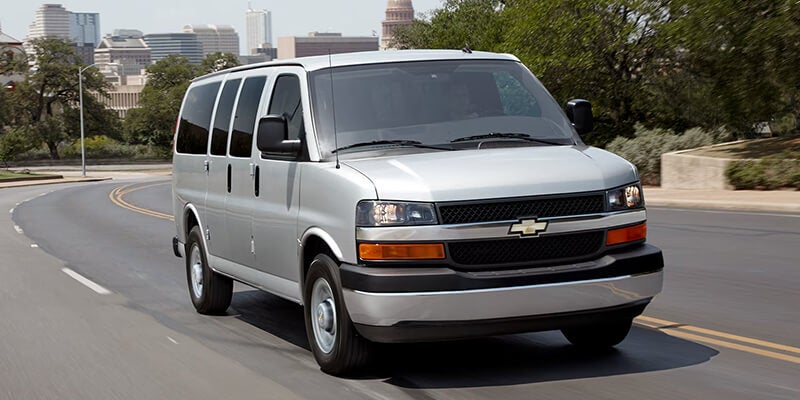 Silver passenger van driving on a paved city road, viewed from the front-left angle