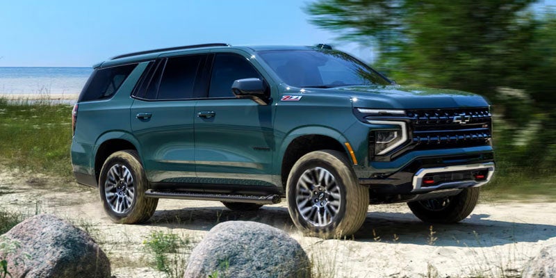 Dark green Chevrolet SUV driving on a sandy path near a lakeside, shown from a front-left angle
